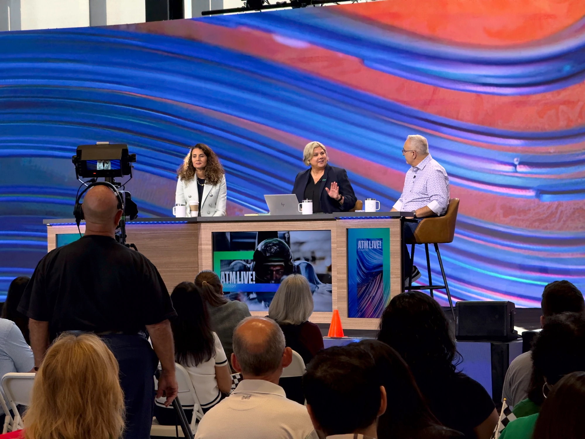 Three panelists live at the branded anchor desk, broadcast camera in the foreground