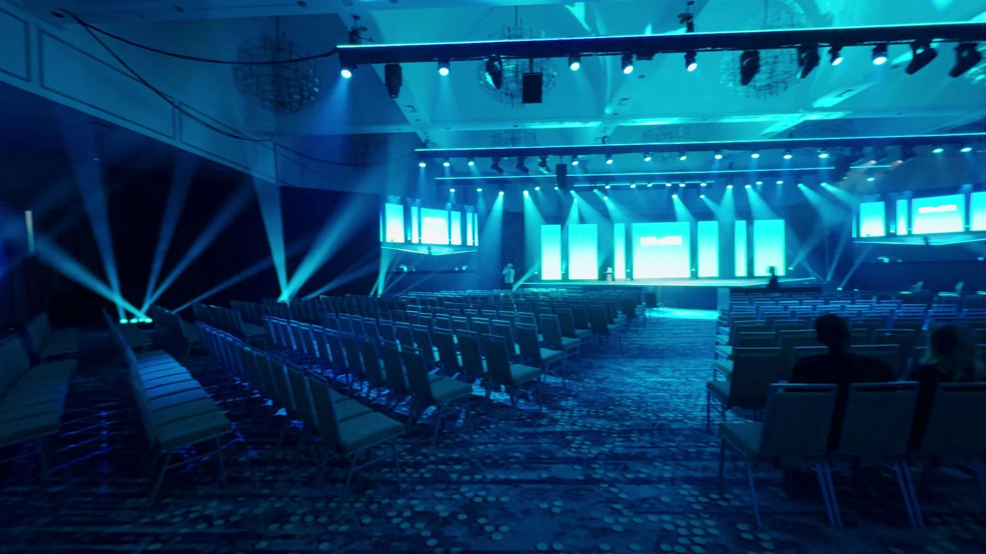 Slow sweep across a finished general session: teal light, beam lighting, and wraparound LED
